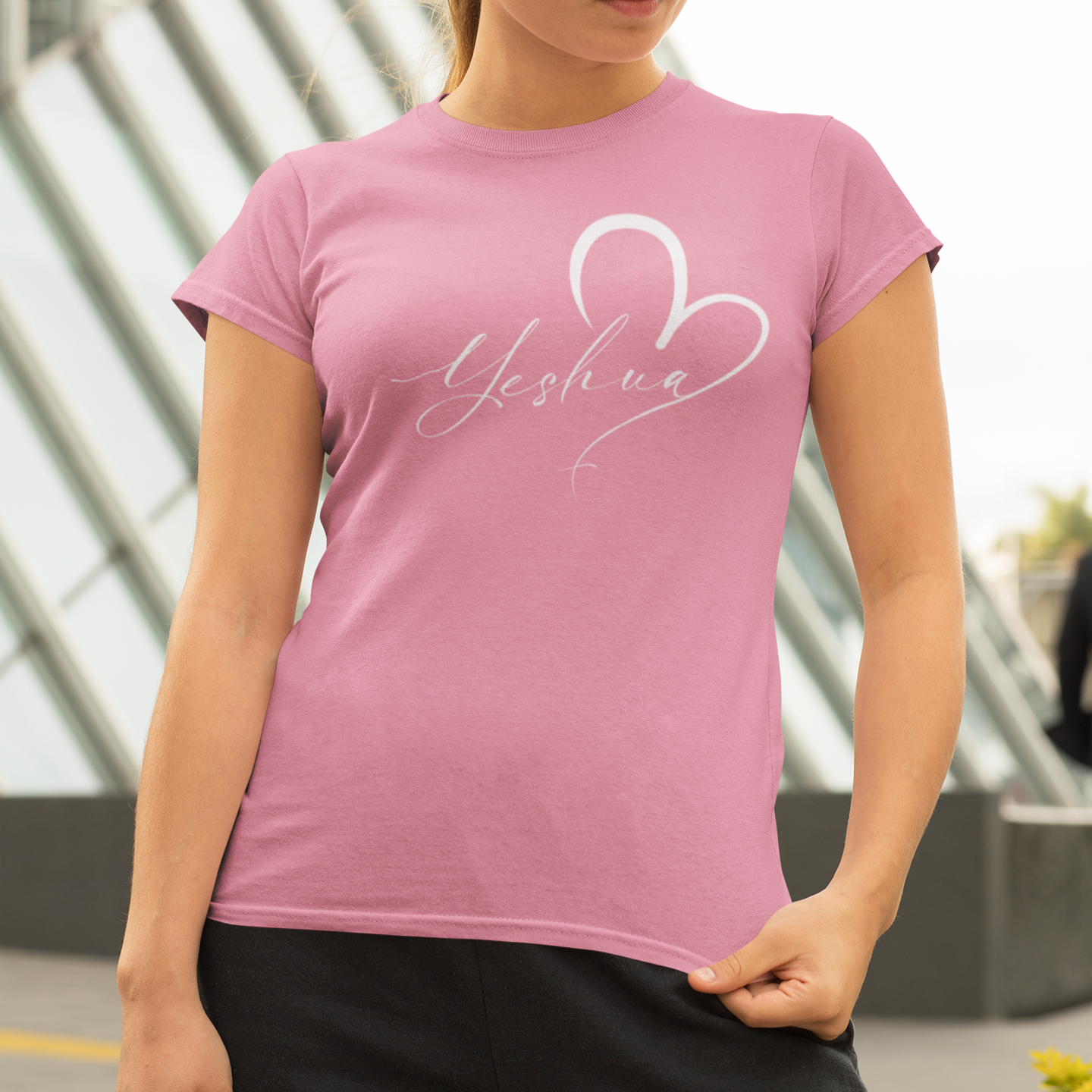 Woman wearing a pink t-shirt with 'Yeshua' design outdoors.