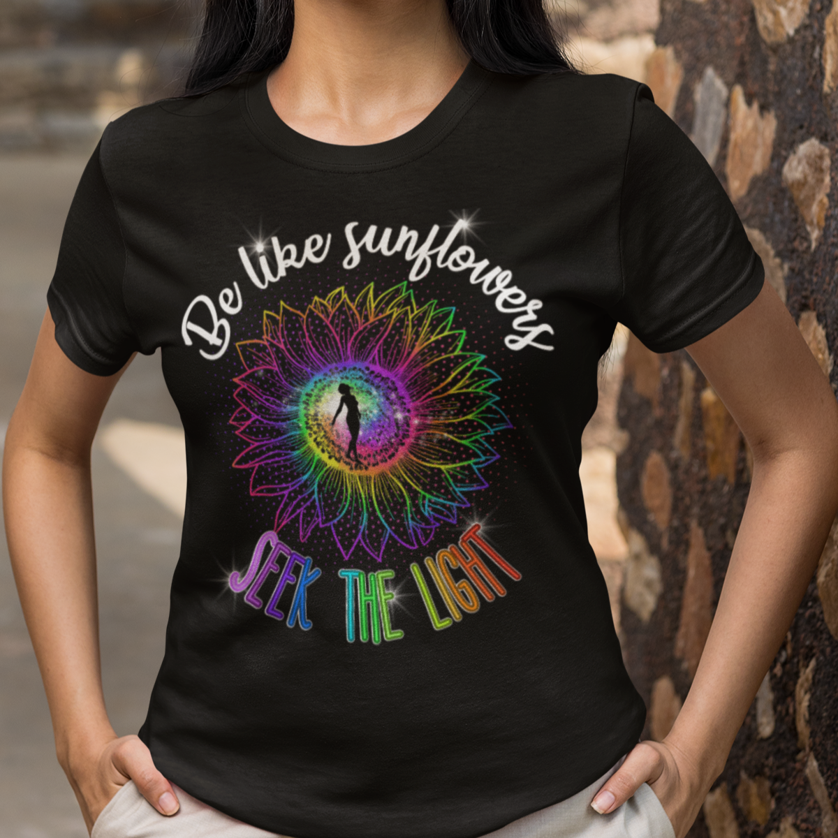Woman wearing a black t-shirt with a colorful design and text, standing against a stone wall.