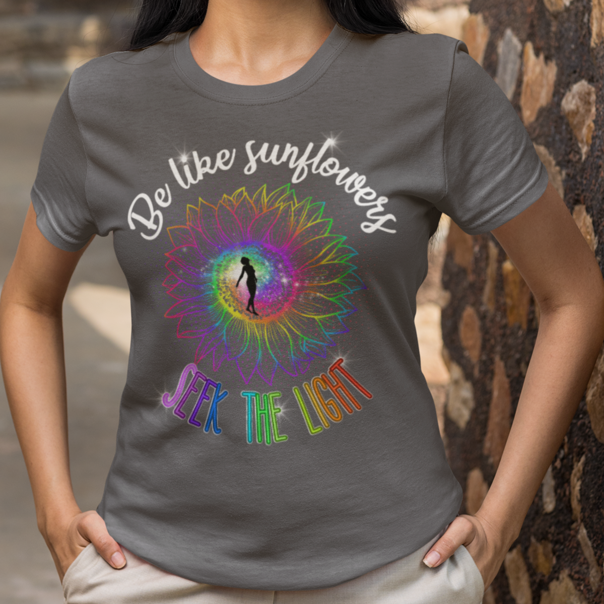Woman wearing a gray t-shirt with a colorful design and text, standing against a stone wall.