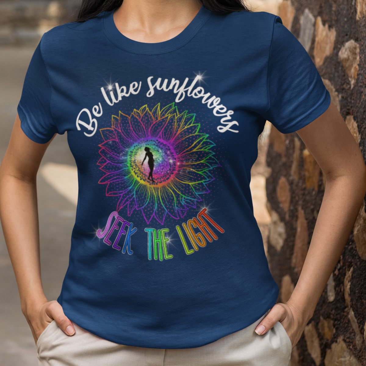 Woman wearing a blue t-shirt with a colorful design and text, standing against a stone wall.