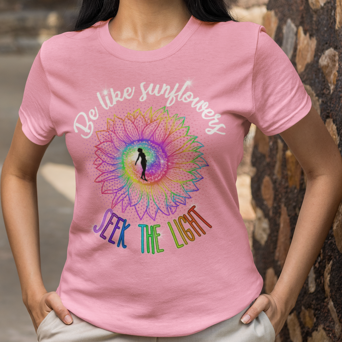 Woman wearing a pink t-shirt with a colorful design and text, standing against a stone wall.
