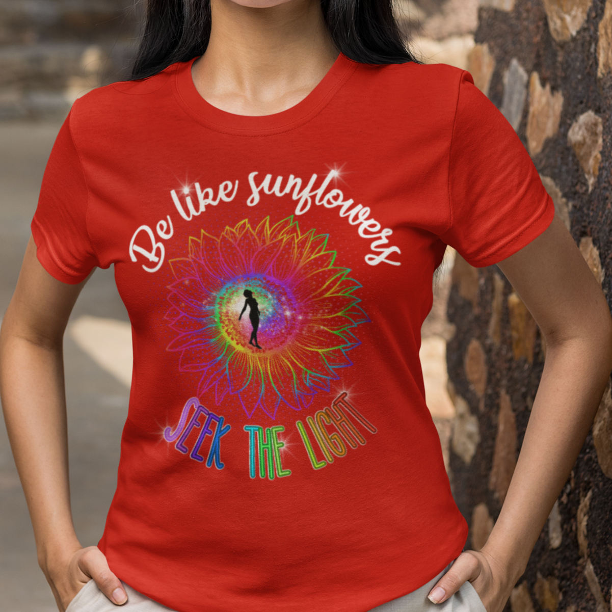 Woman wearing a red t-shirt with a colorful design and text, standing against a stone wall.