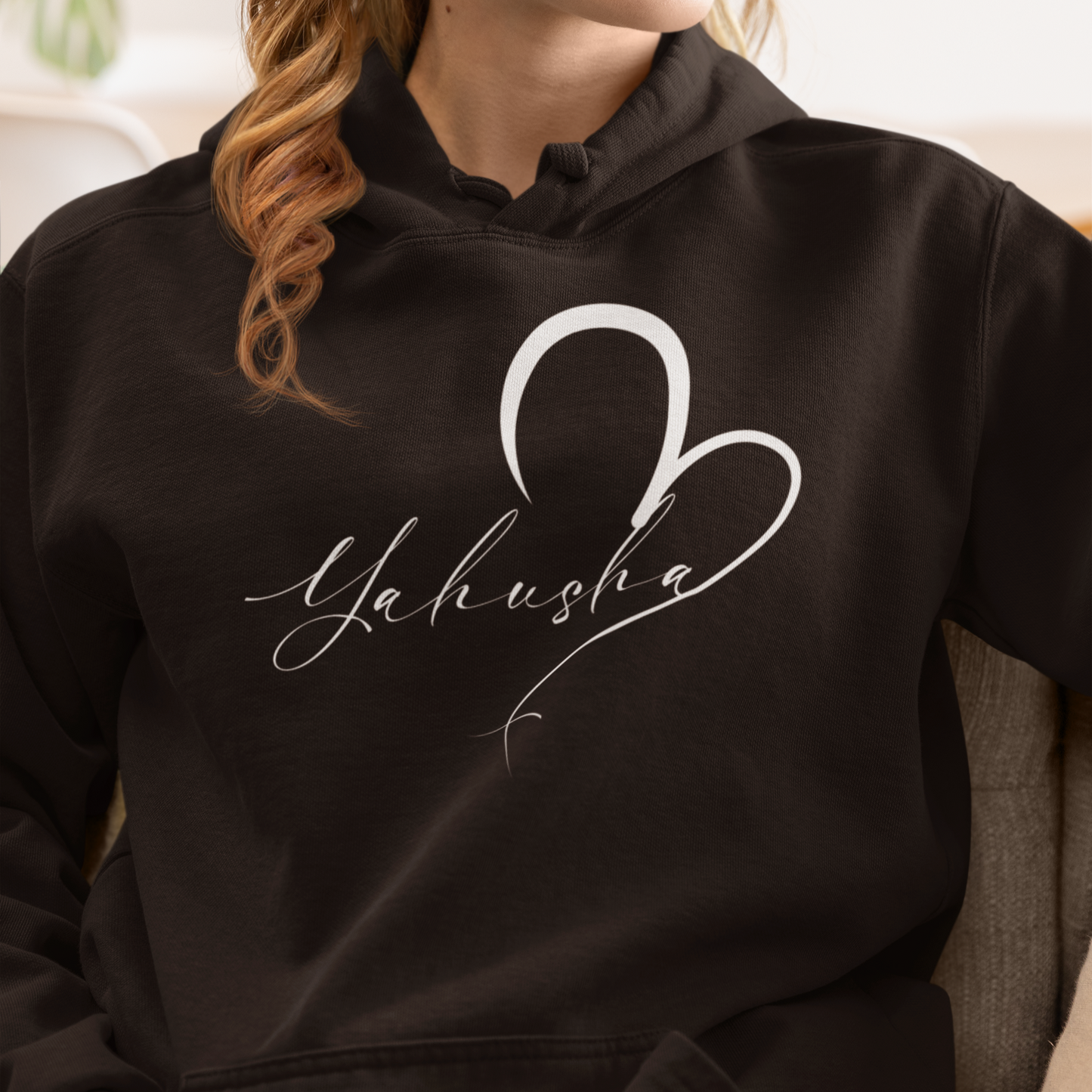 Person wearing a black hoodie with 'Yahusha' design, sitting on a couch.