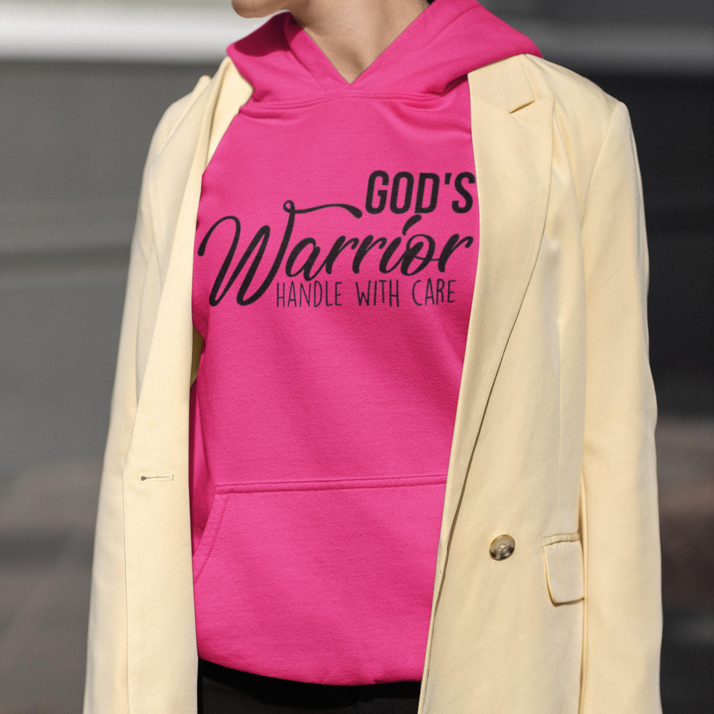 Person wearing a pink hoodie with 'God's Warrior Handle with Care' text and a beige blazer.