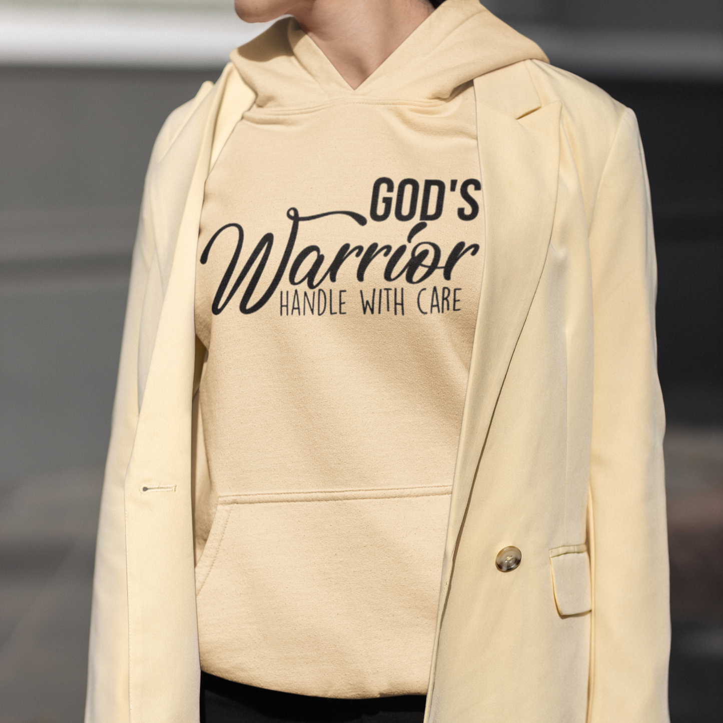 Beige hoodie with 'God's Warrior Handle with Care' text worn by a person.