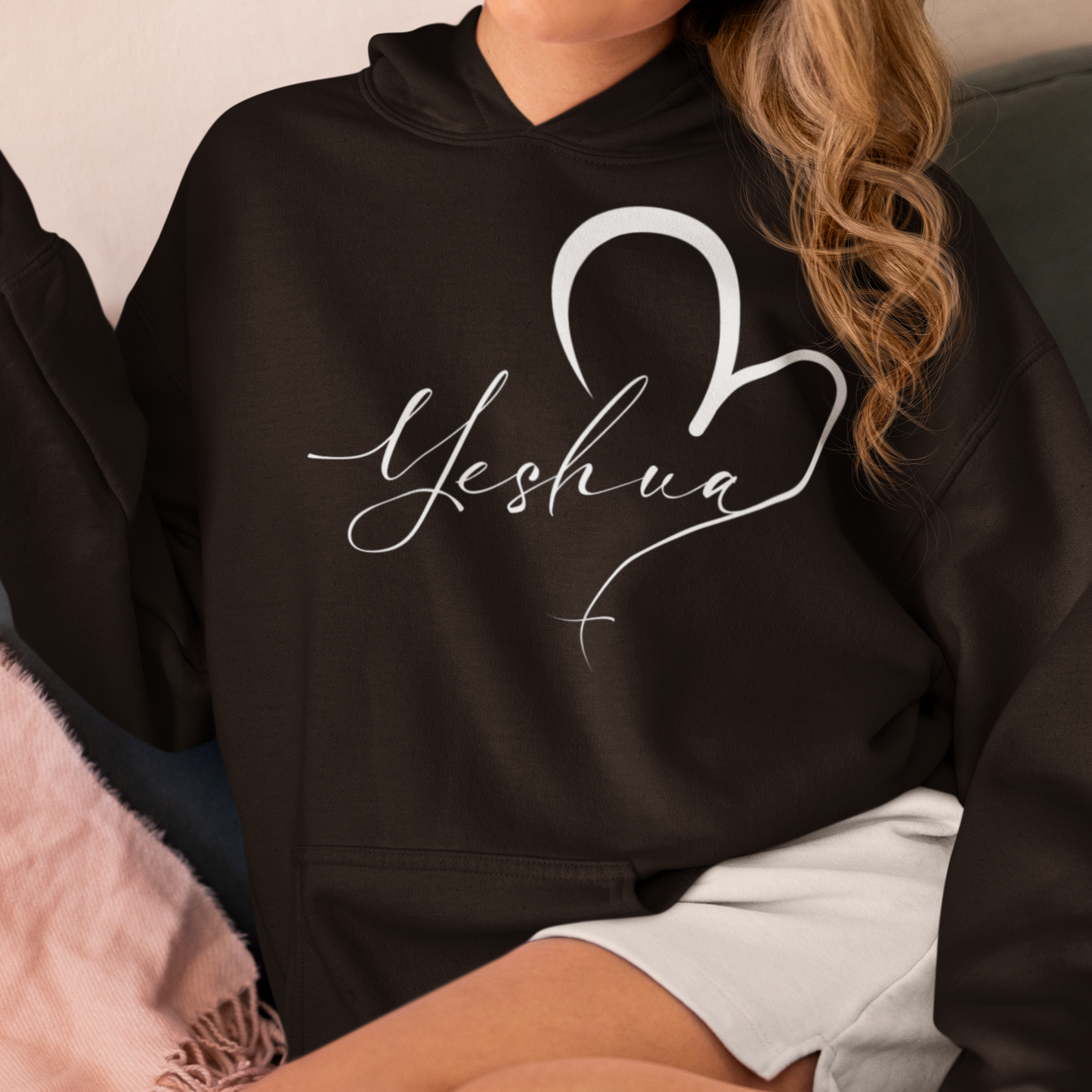 Person wearing a black hoodie with 'Yeshua' and a heart design, sitting on a couch.
