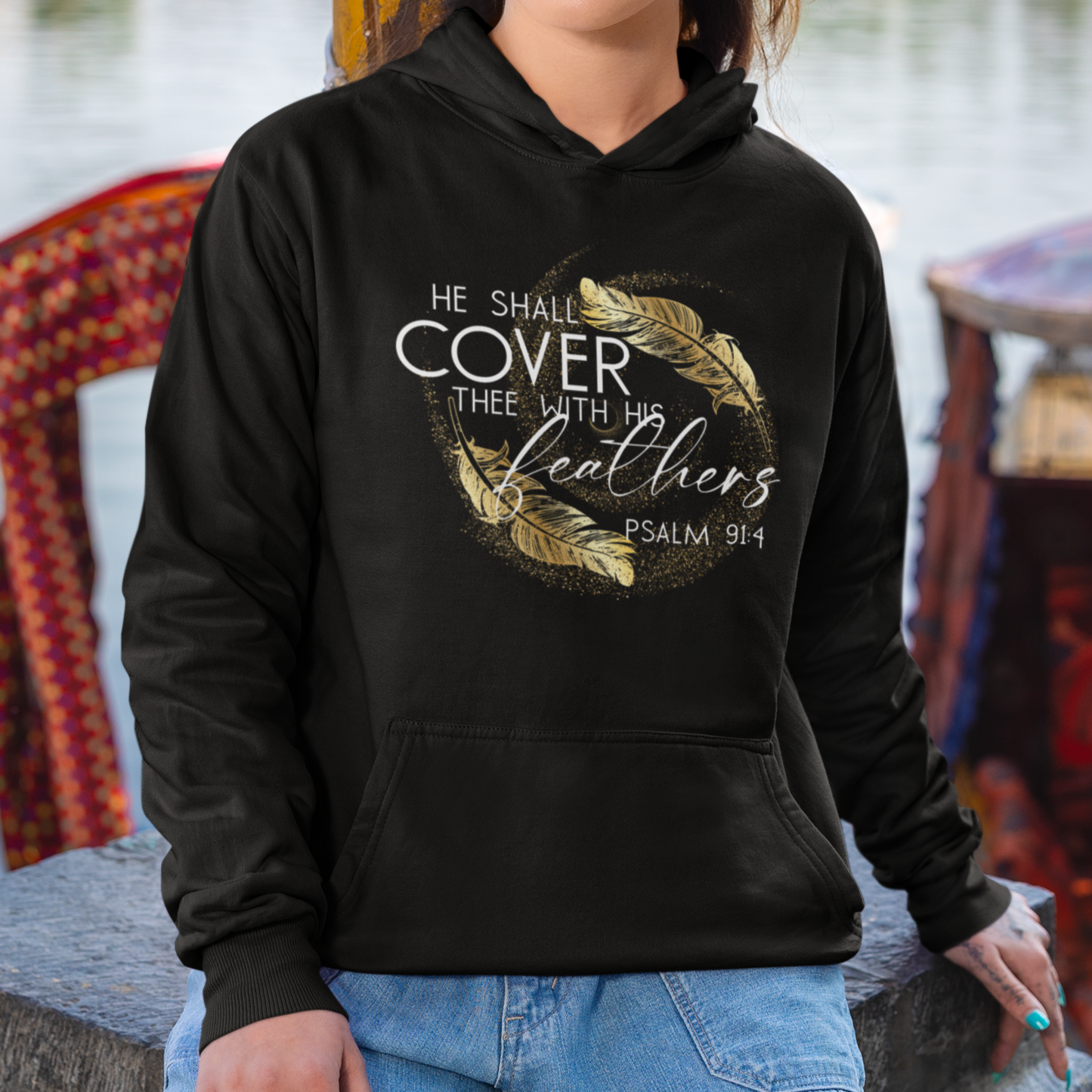 Woman wearing a black hoodie with text and gold design, standing by a waterfront with colorful boats and umbrellas.