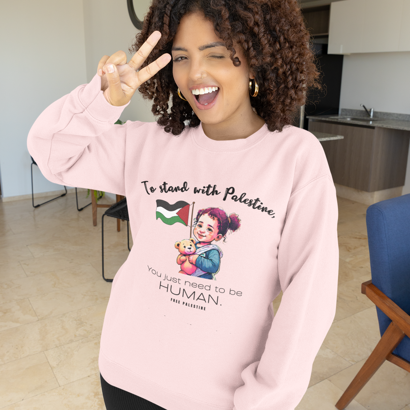 Person wearing a pink sweatshirt with a graphic design and text, making a peace sign indoors.