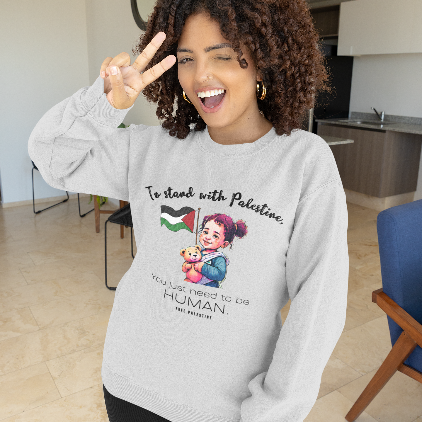 Woman wearing a gray sweatshirt with a graphic design and text, making a peace sign indoors.