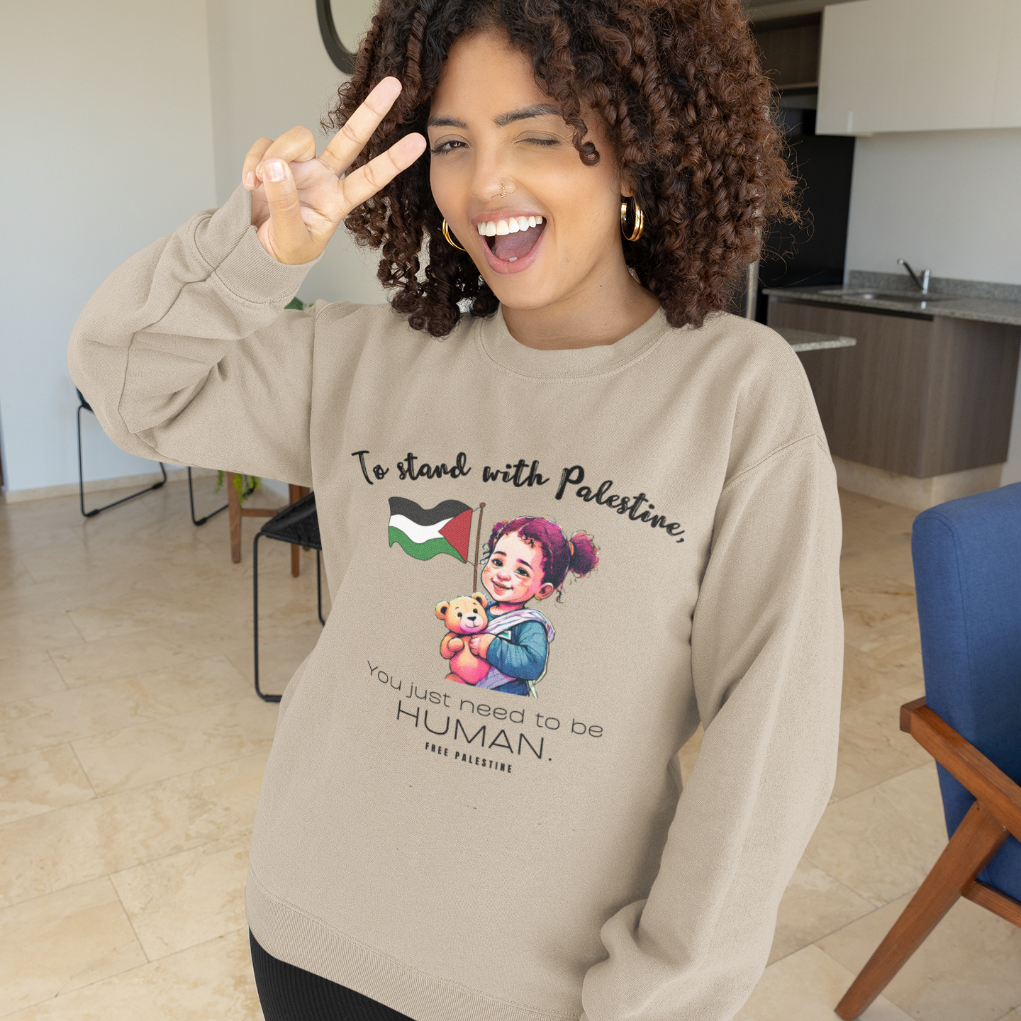 Person wearing a beige sweatshirt with a graphic and text, making a peace sign indoors.