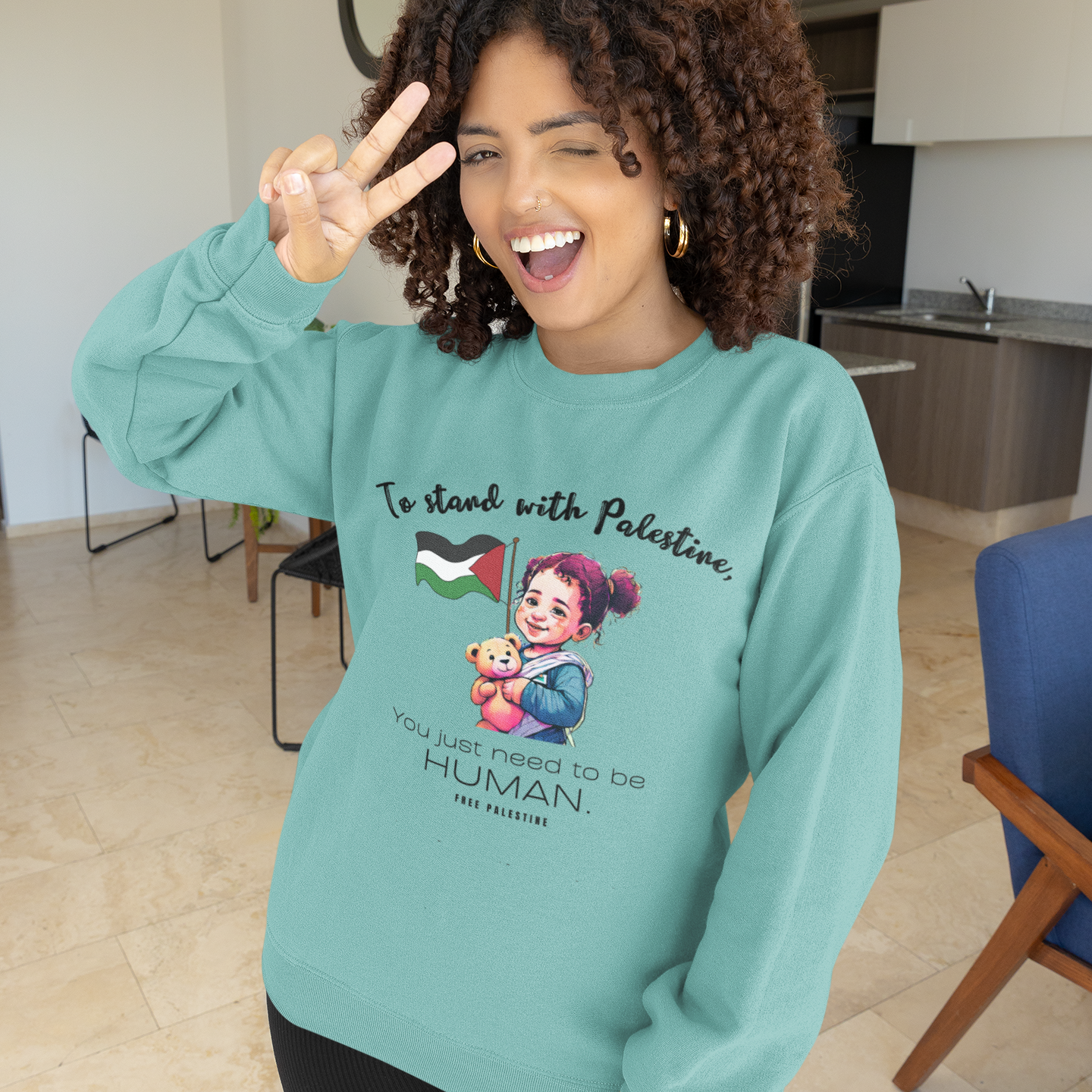 Woman wearing a teal sweatshirt with a graphic design and text, standing in a modern indoor setting.