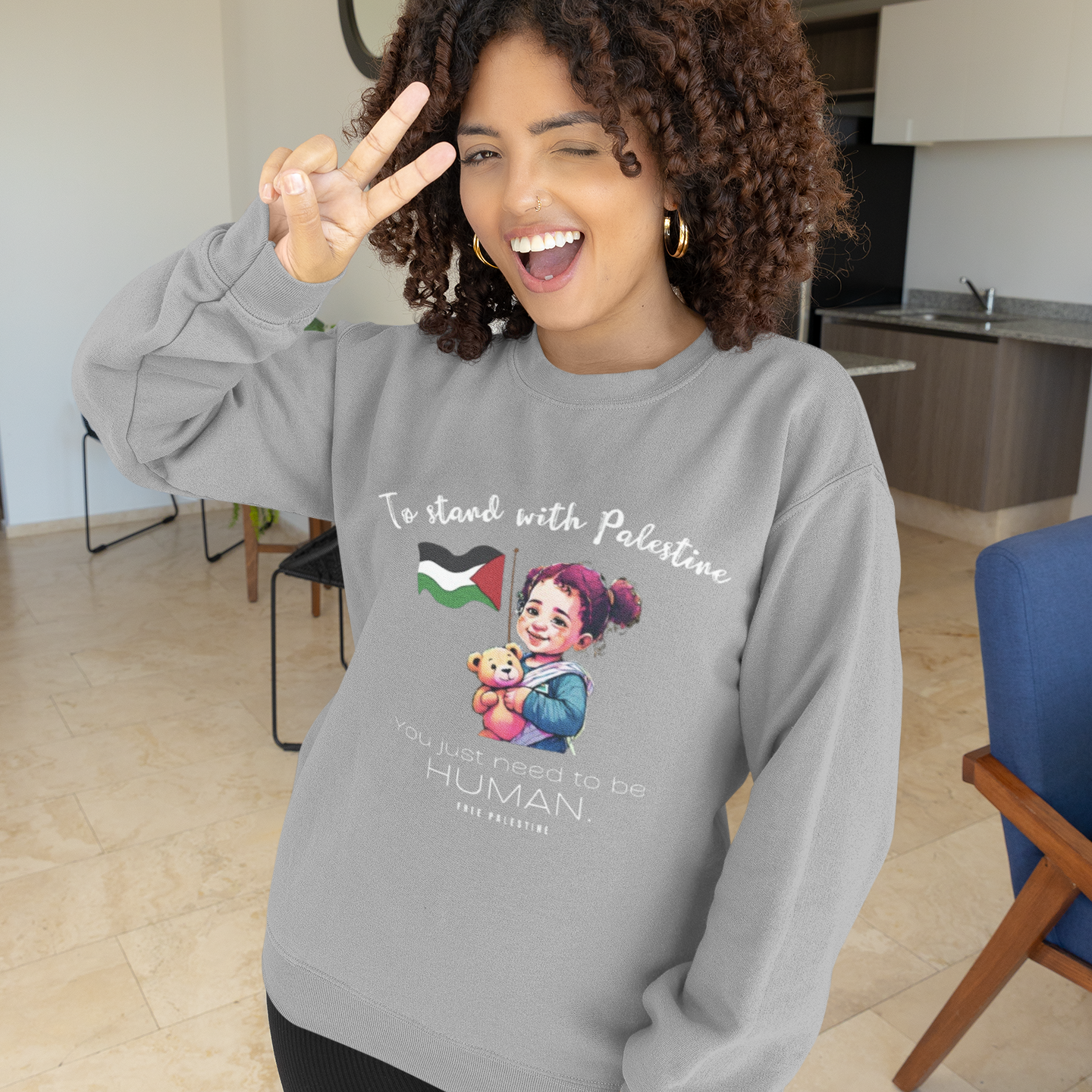 Woman wearing a gray sweatshirt with a graphic and text, making a peace sign indoors.