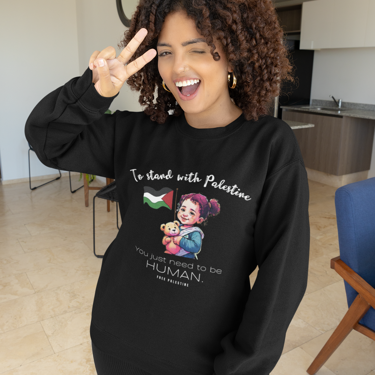 Woman wearing a black sweatshirt with a graphic and text, making a peace sign indoors.