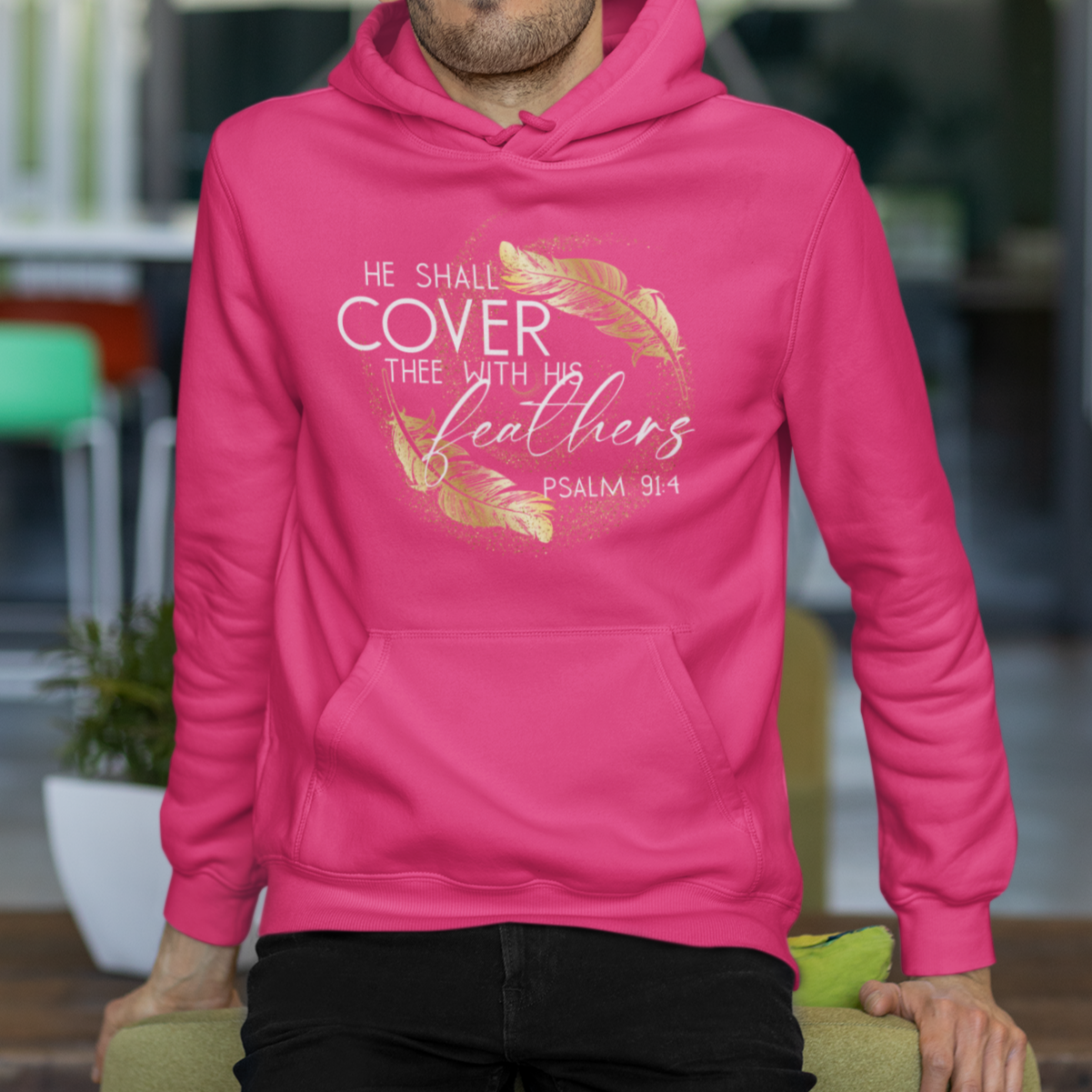 Man wearing a pink hoodie with text and feather design, sitting on a chair indoors.