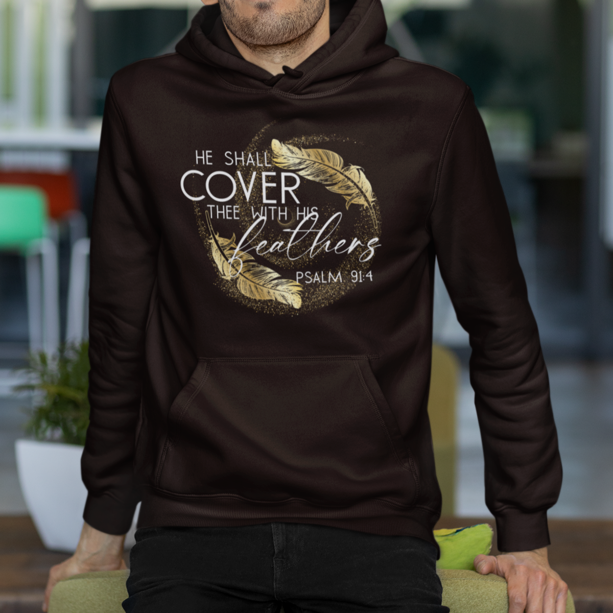 Man wearing a brown hoodie with text and feather design, standing in an outdoor setting.