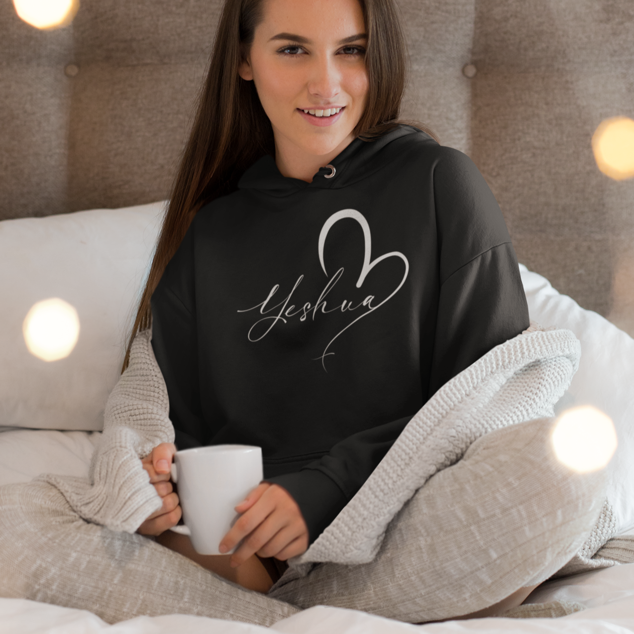 Woman wearing a black hoodie with 'Yeshua' design, sitting on a bed with lights in the background