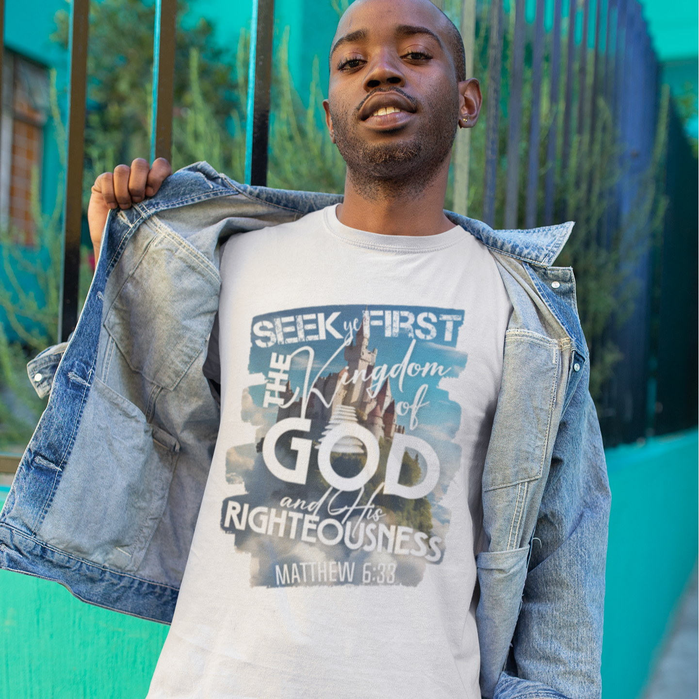 Man wearing a white t-shirt with a religious quote, standing against a teal wall.