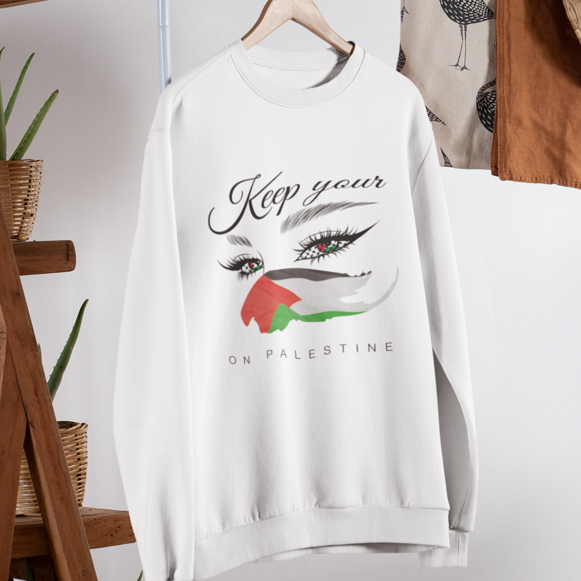 White sweatshirt with graphic design and text on a rack against a white wall with wooden shelves and plants.