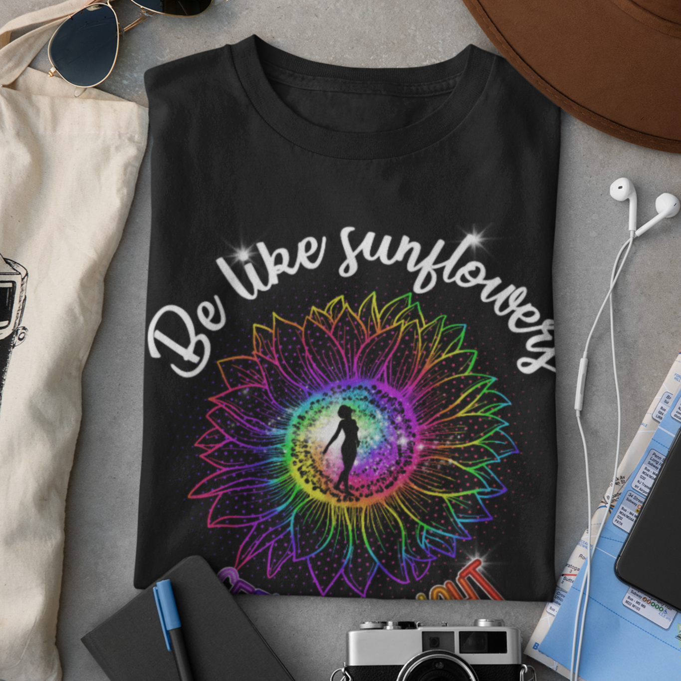 Black t-shirt with colorful sunflower design and text 'Be like sunflowers' on a gray surface with accessories.
