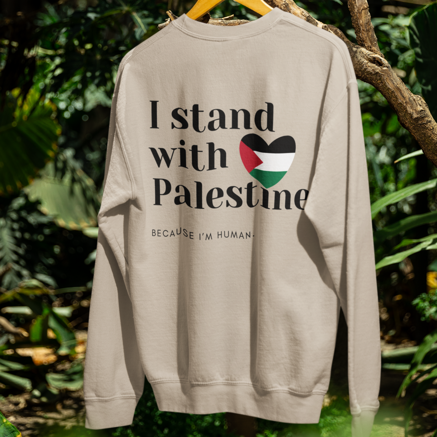Beige sweatshirt with 'I stand with Palestine because I'm human' text and Palestinian flag design, hanging on a tree branch with green foliage in the background.