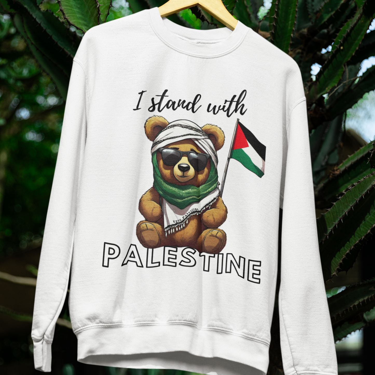 White sweatshirt with a bear graphic and 'I stand with Palestine' text, hanging against a green leafy background.