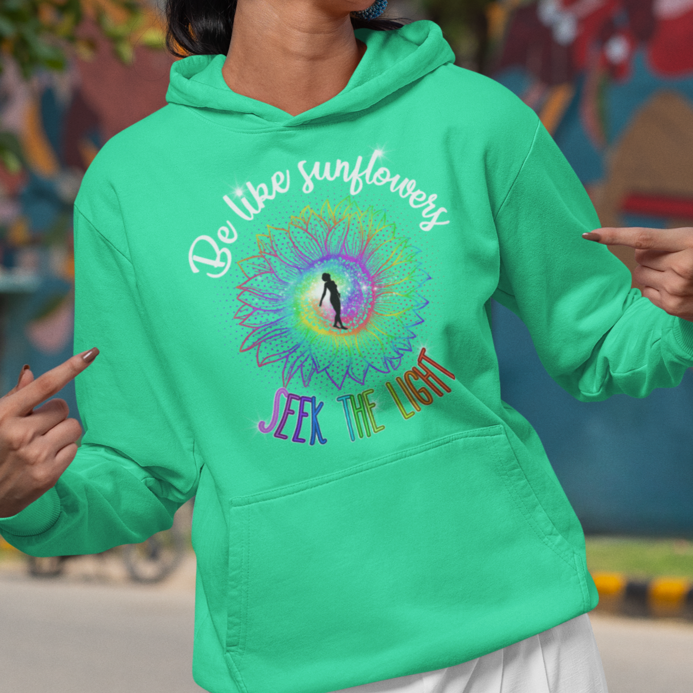 Woman wearing a green hoodie with text and graphics, standing in front of a colorful mural.
