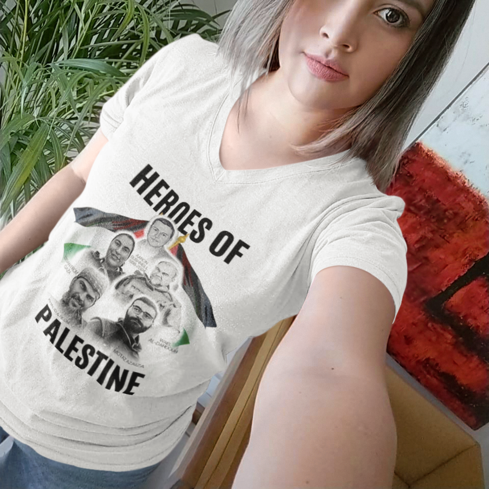 Person wearing a white t-shirt with 'Heroes of Palestine' design, sitting indoors with plants and a painting in the background.
