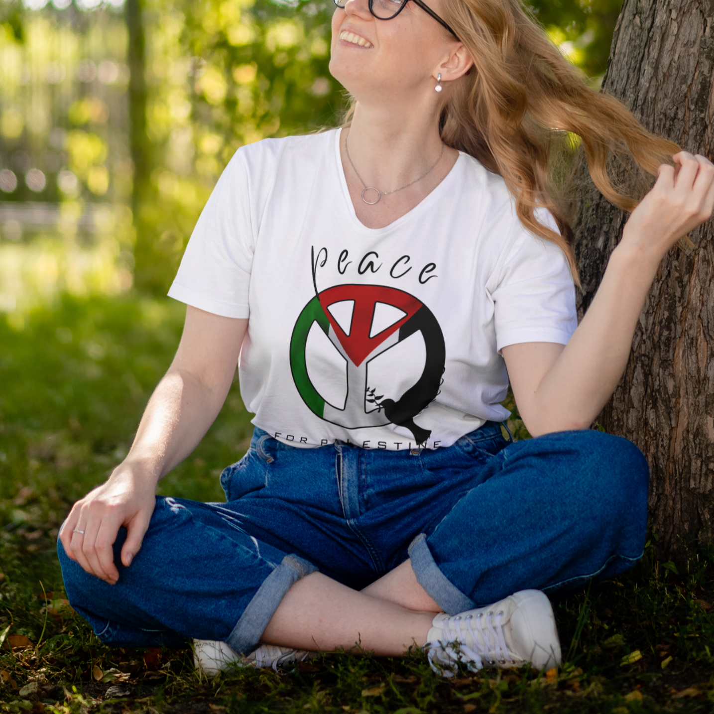 Person sitting under a tree wearing a white t-shirt with a peace symbol design.