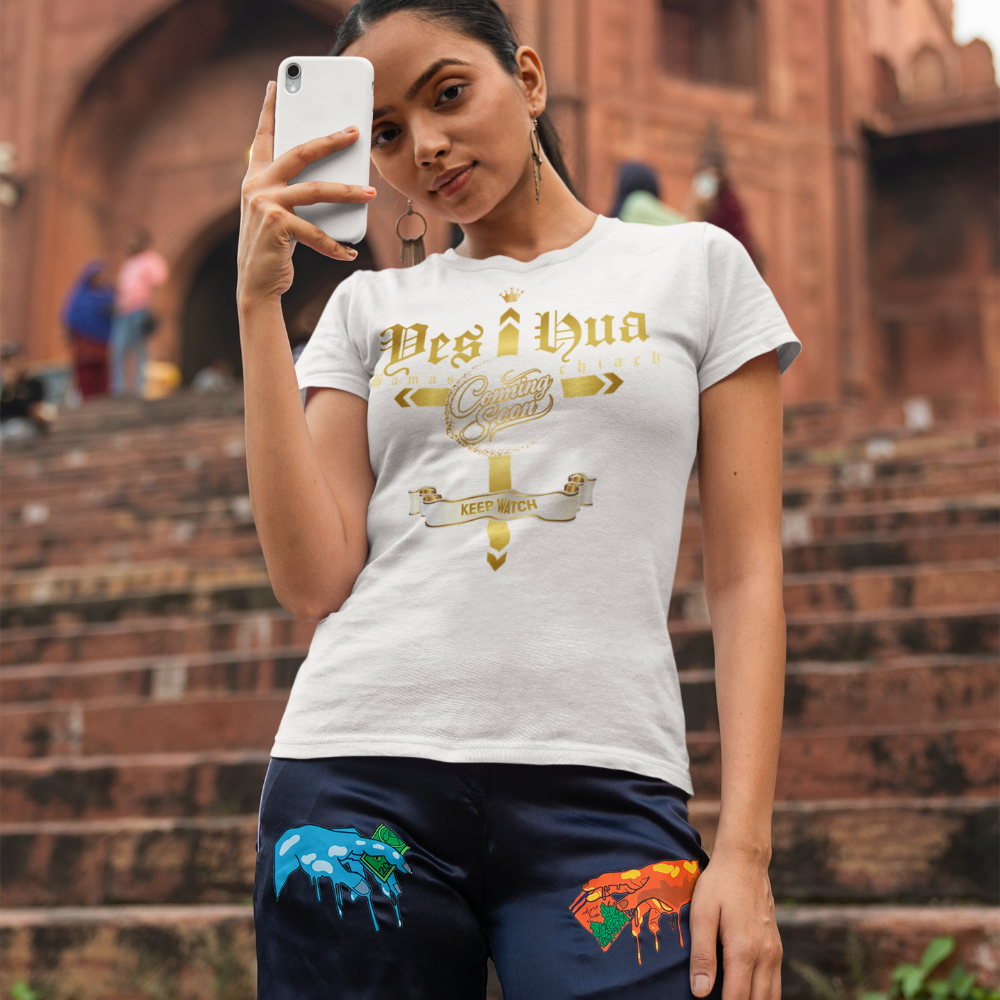 Woman taking a selfie wearing a white t-shirt with gold text and navy pants with colorful designs, standing on steps in front of a brick building.