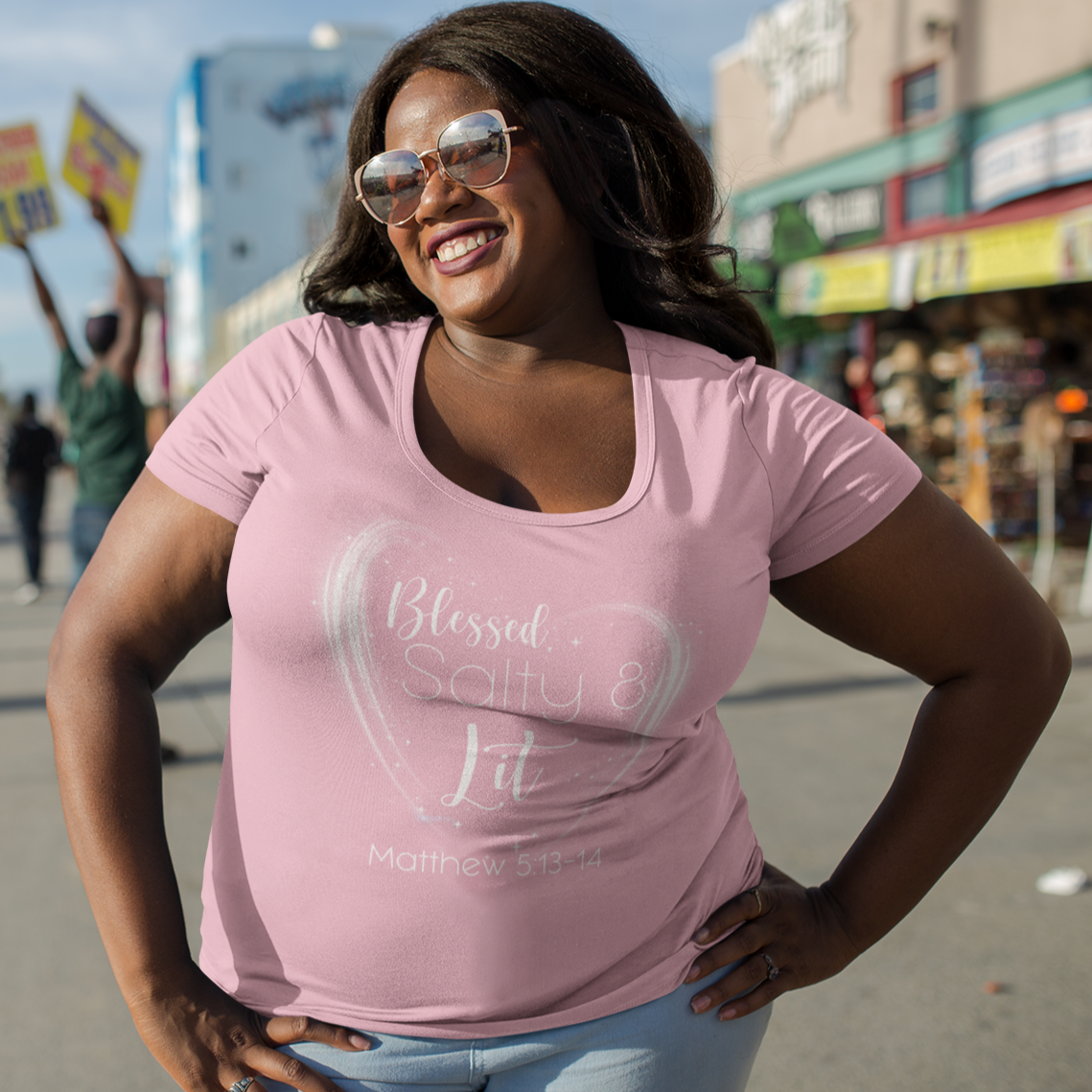 Woman wearing a pink t-shirt with text, standing on a street with palm trees and buildings in the background.