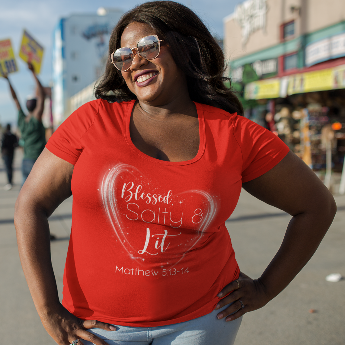 Woman wearing a red t-shirt with a heart design and text, standing on a street with palm trees and buildings in the background.