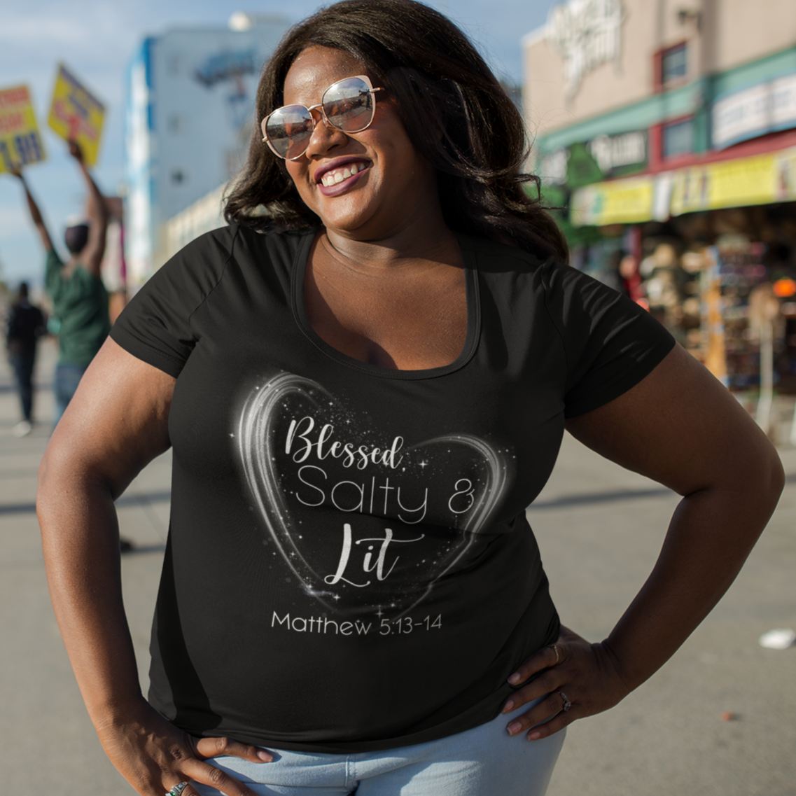 Woman wearing a black t-shirt with text, standing on a street with palm trees and buildings in the background.
