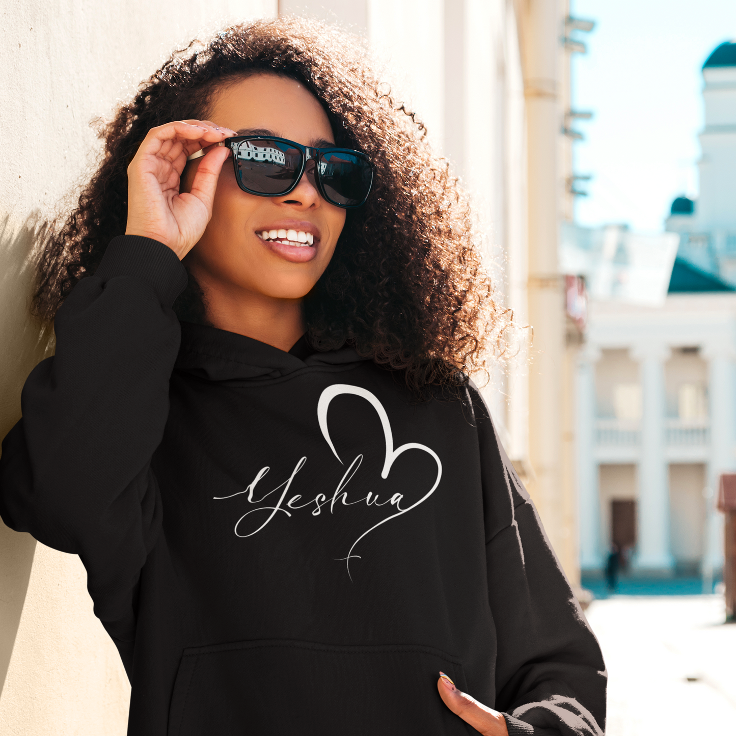 Person wearing a black hoodie with 'Yeshua' design, holding sunglasses, outdoors.