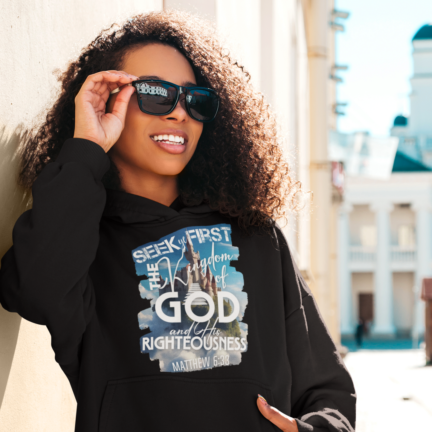 Person wearing a black hoodie with a religious text design, holding sunglasses.