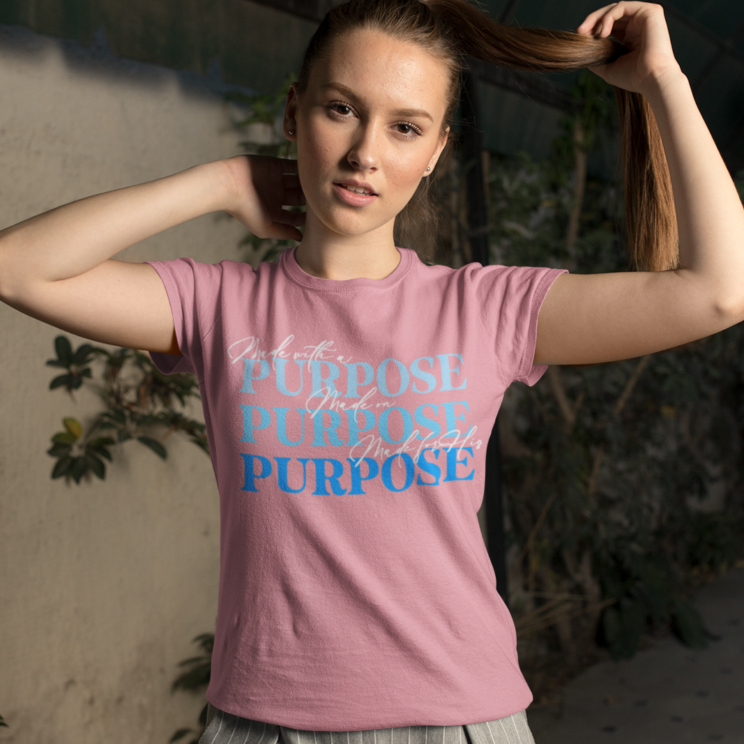 Woman wearing a pink t-shirt with 'PURPOSE' printed on it, standing indoors with plants around.