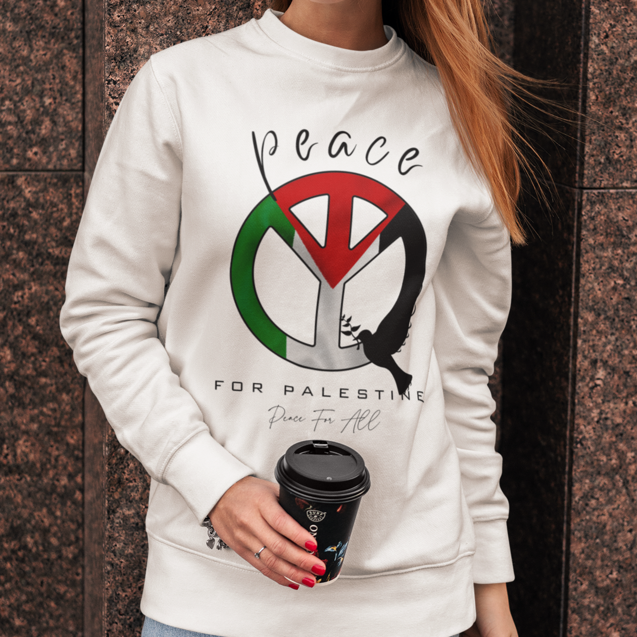 Woman wearing a white sweatshirt with a peace symbol design, holding a black cup against a brown wall.