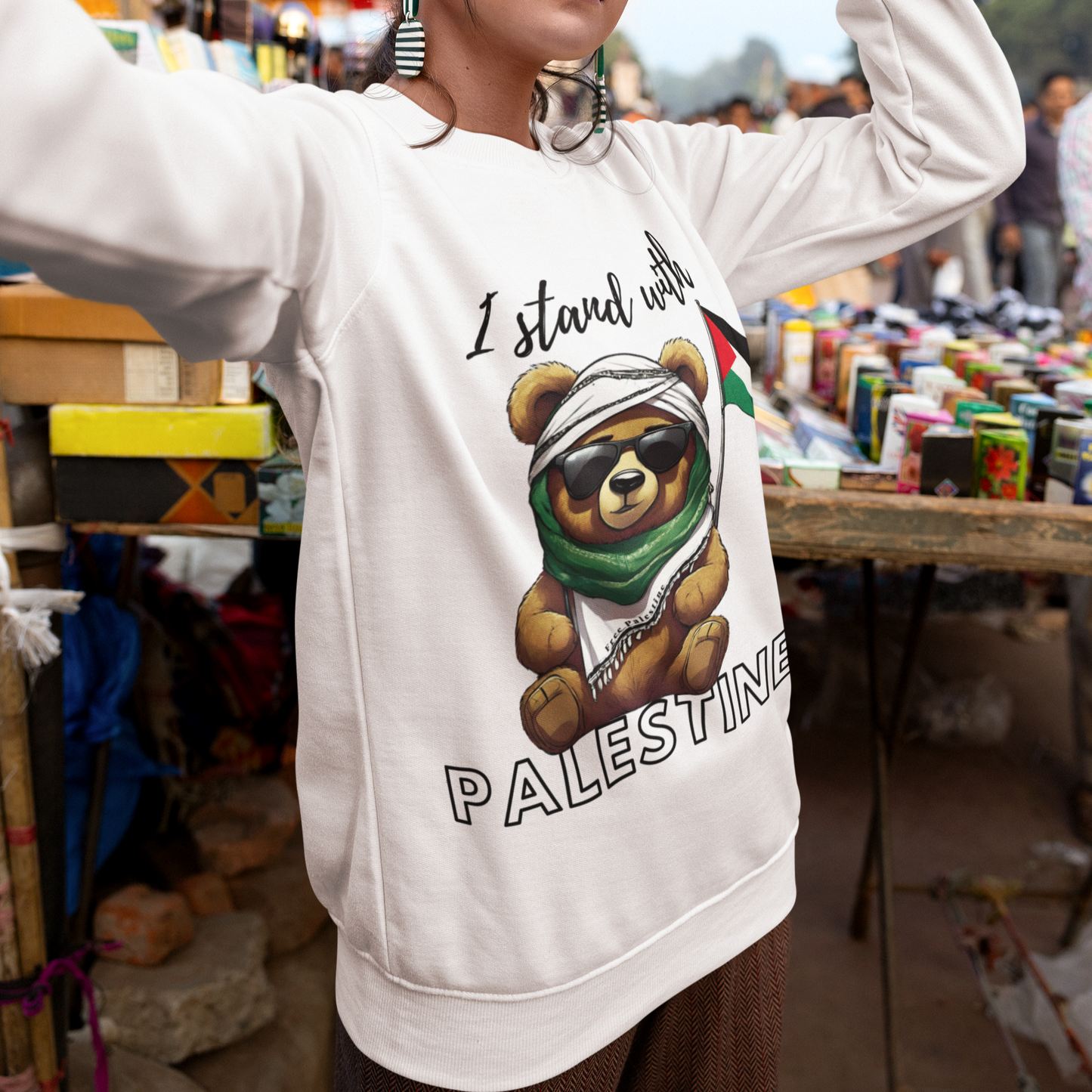 Person wearing a white sweatshirt with a bear graphic and 'I stand with Palestine' text in an outdoor market setting.