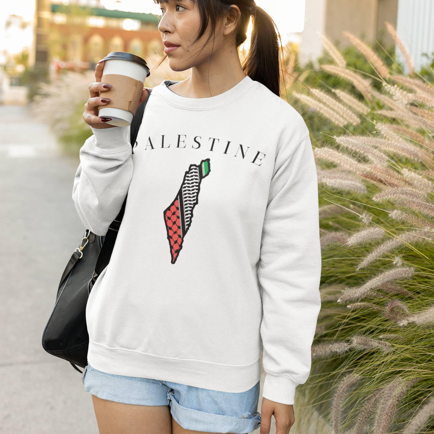 Woman wearing a white sweatshirt with a design and text, holding a coffee cup outdoors.