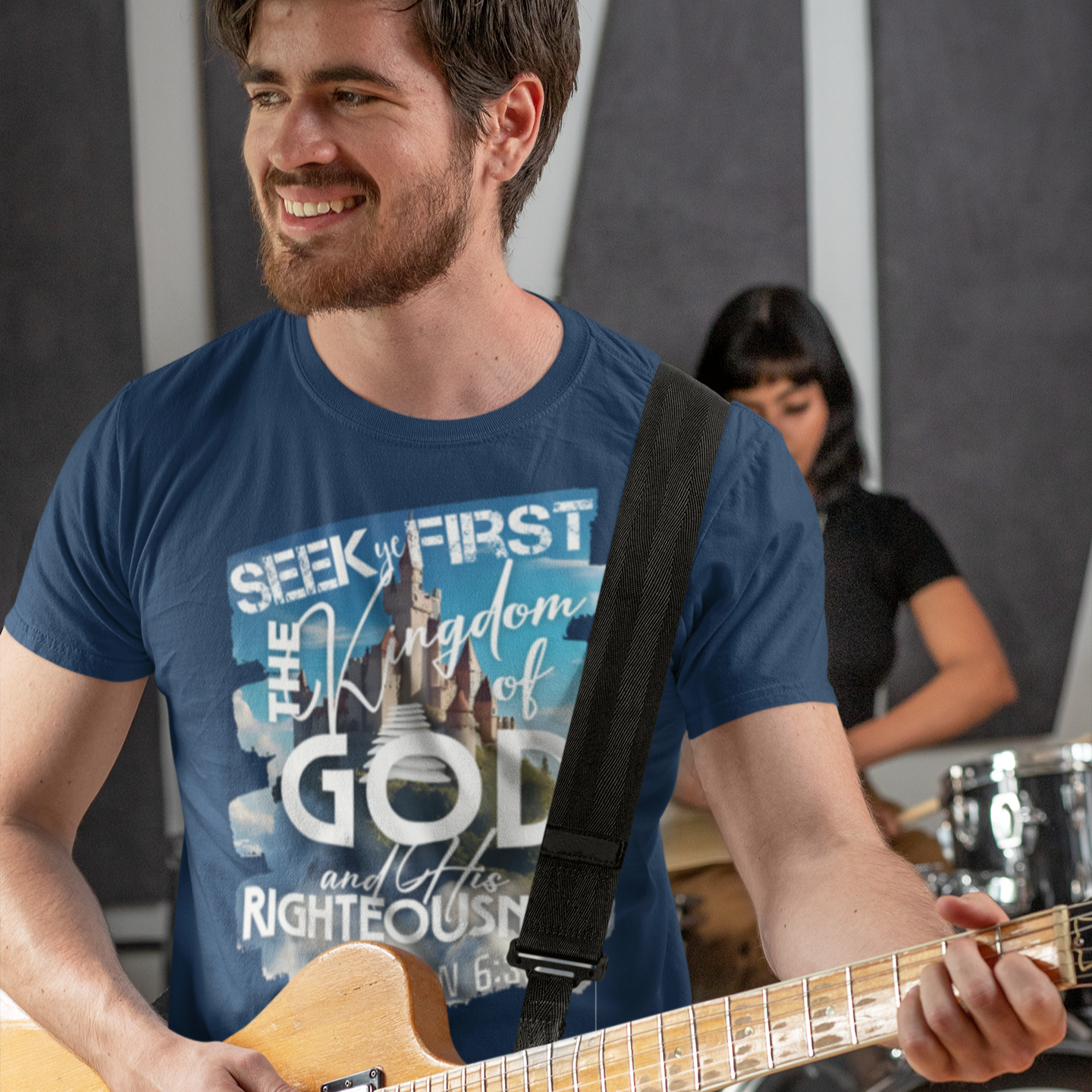Man playing guitar wearing a blue t-shirt with a bible verse, with a blurred background of another person and musical instruments.