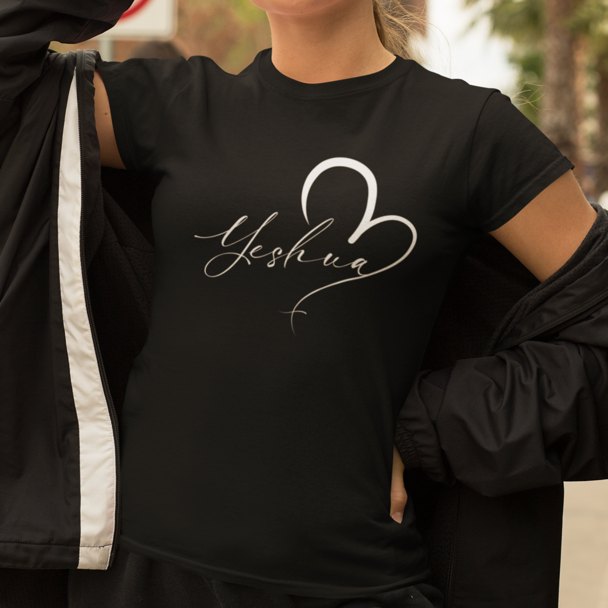 Person wearing a black t-shirt with 'Yeshua' design, holding sunglasses, outdoors.