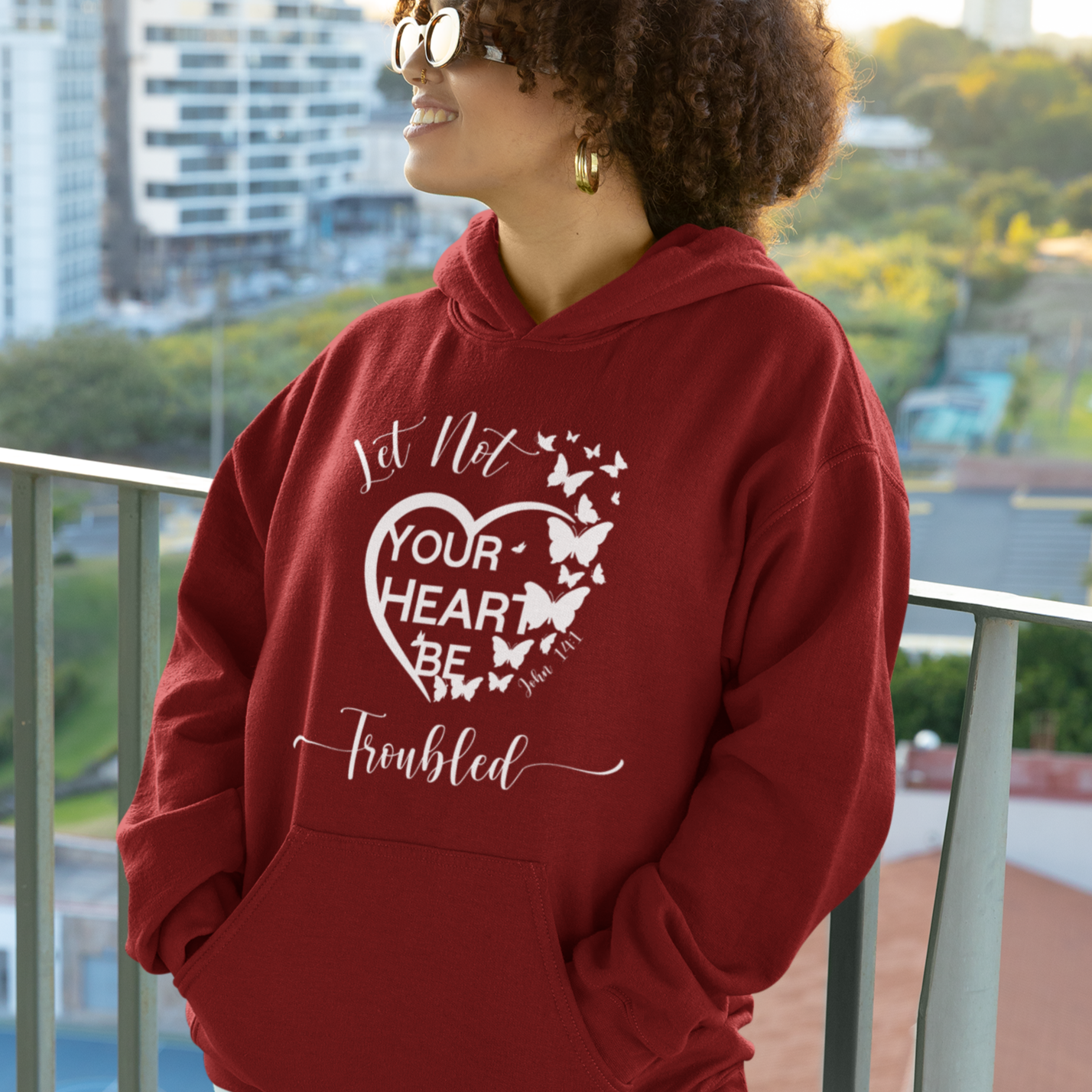 Person wearing a red hoodie with a heart design and text, standing on a balcony with a cityscape background.