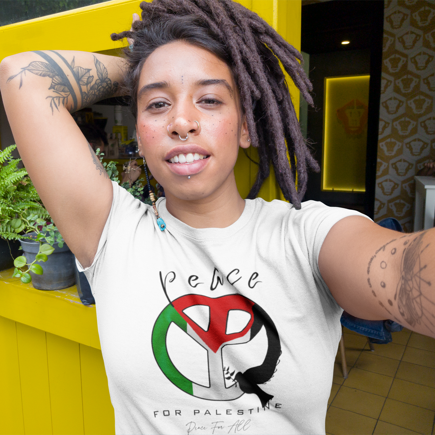 Person wearing a white t-shirt with a peace symbol design, sitting against a yellow wall with plants.