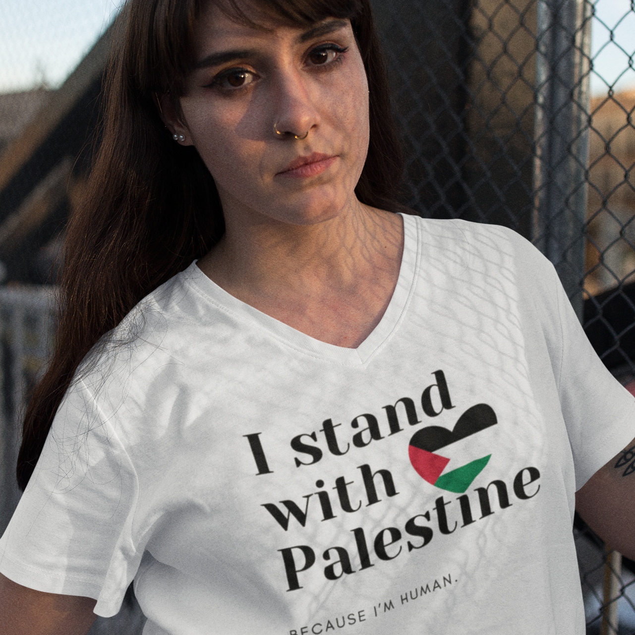 Person wearing a white t-shirt with 'I stand with Palestine' text, standing in front of a chain-link fence.