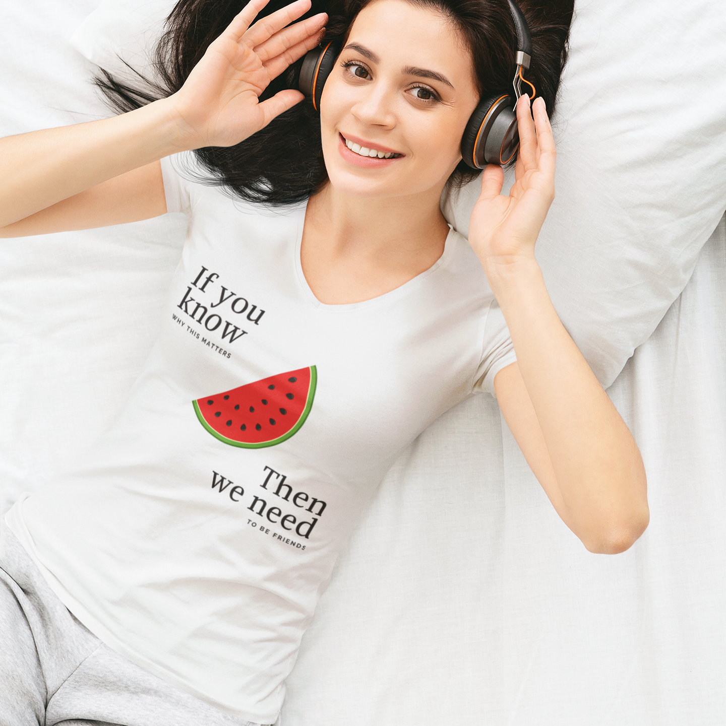 Woman wearing a white t-shirt with text and a watermelon graphic, lying on a bed with headphones.