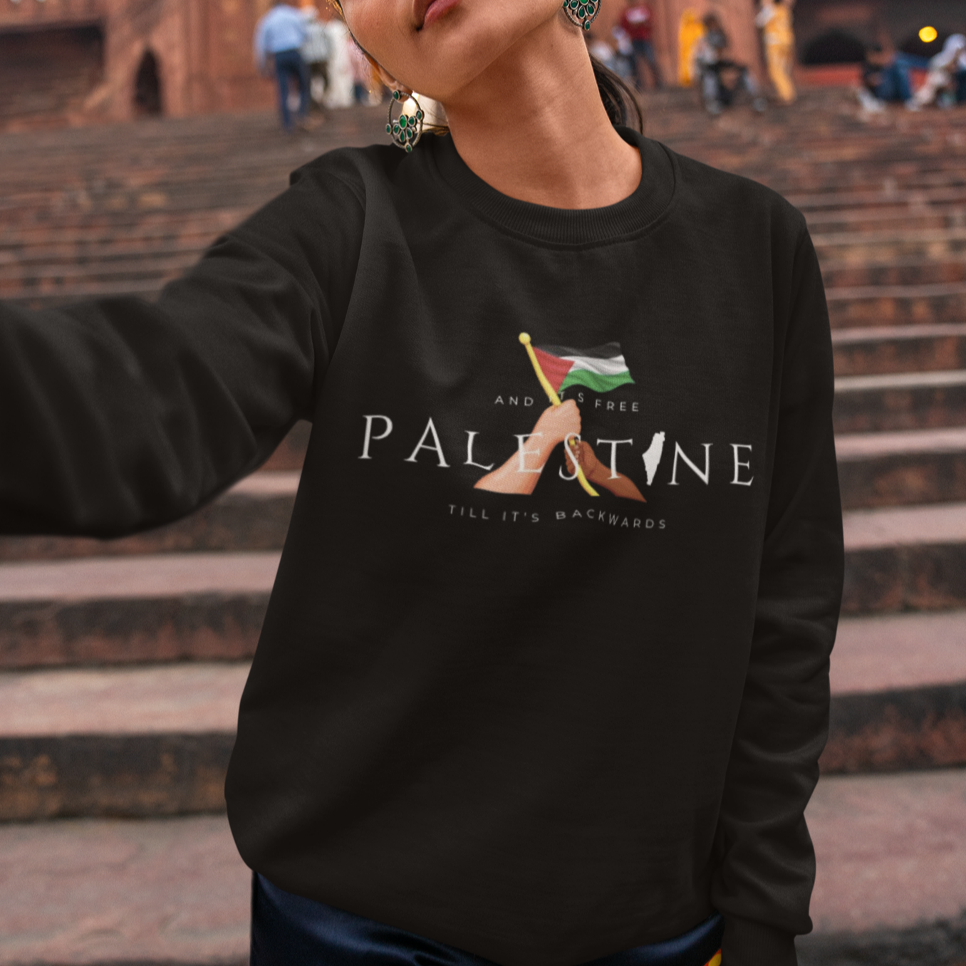 Person wearing a black sweatshirt with 'Palestine' text and flag design, standing on steps in front of a historic building.