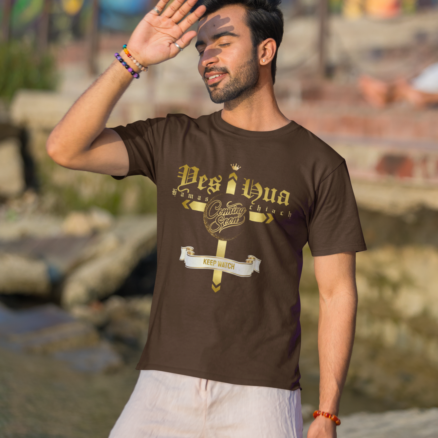 Man wearing a brown t-shirt with text, standing outdoors near a body of water with colorful background.