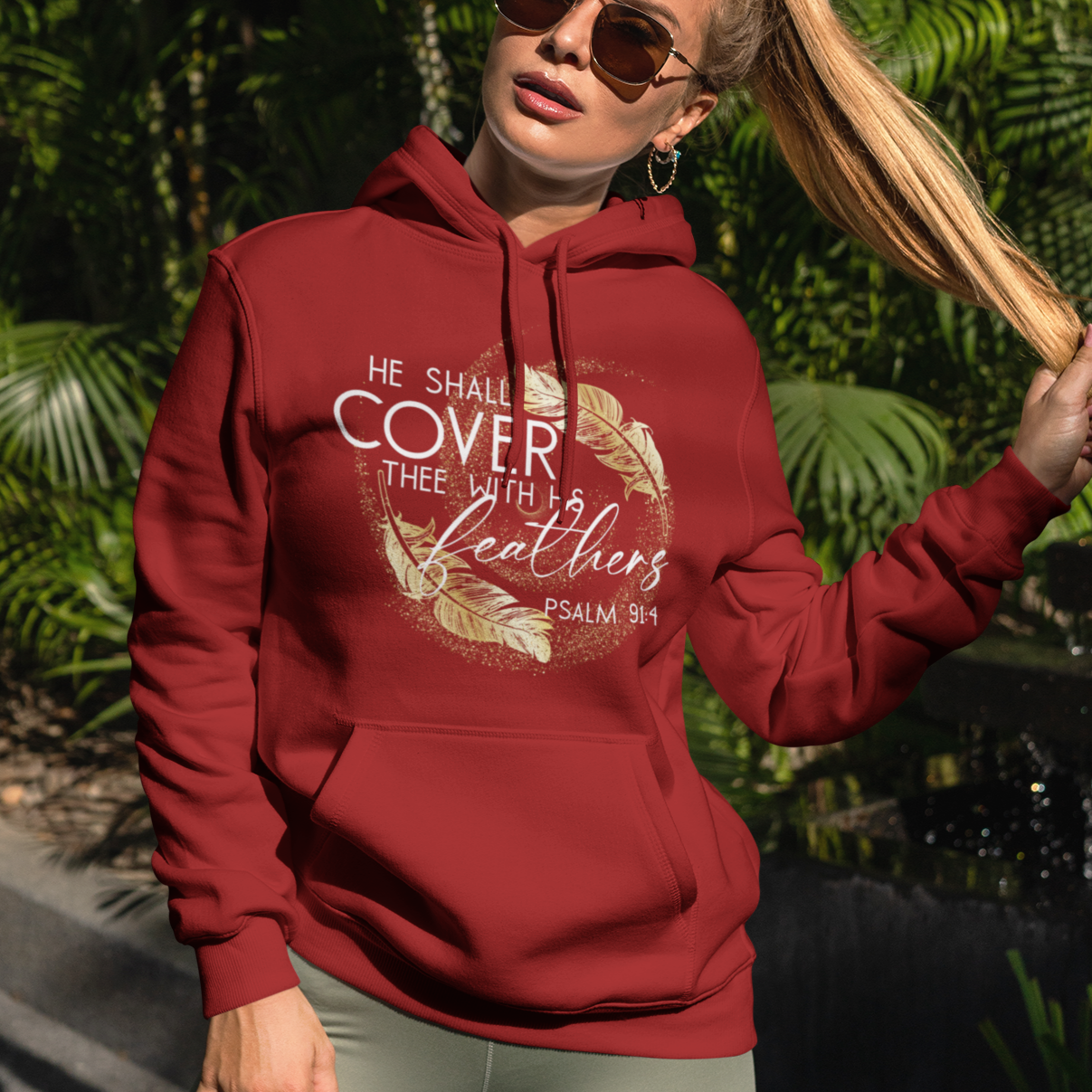 Woman wearing a red hoodie with text and design, standing outdoors with greenery in the background