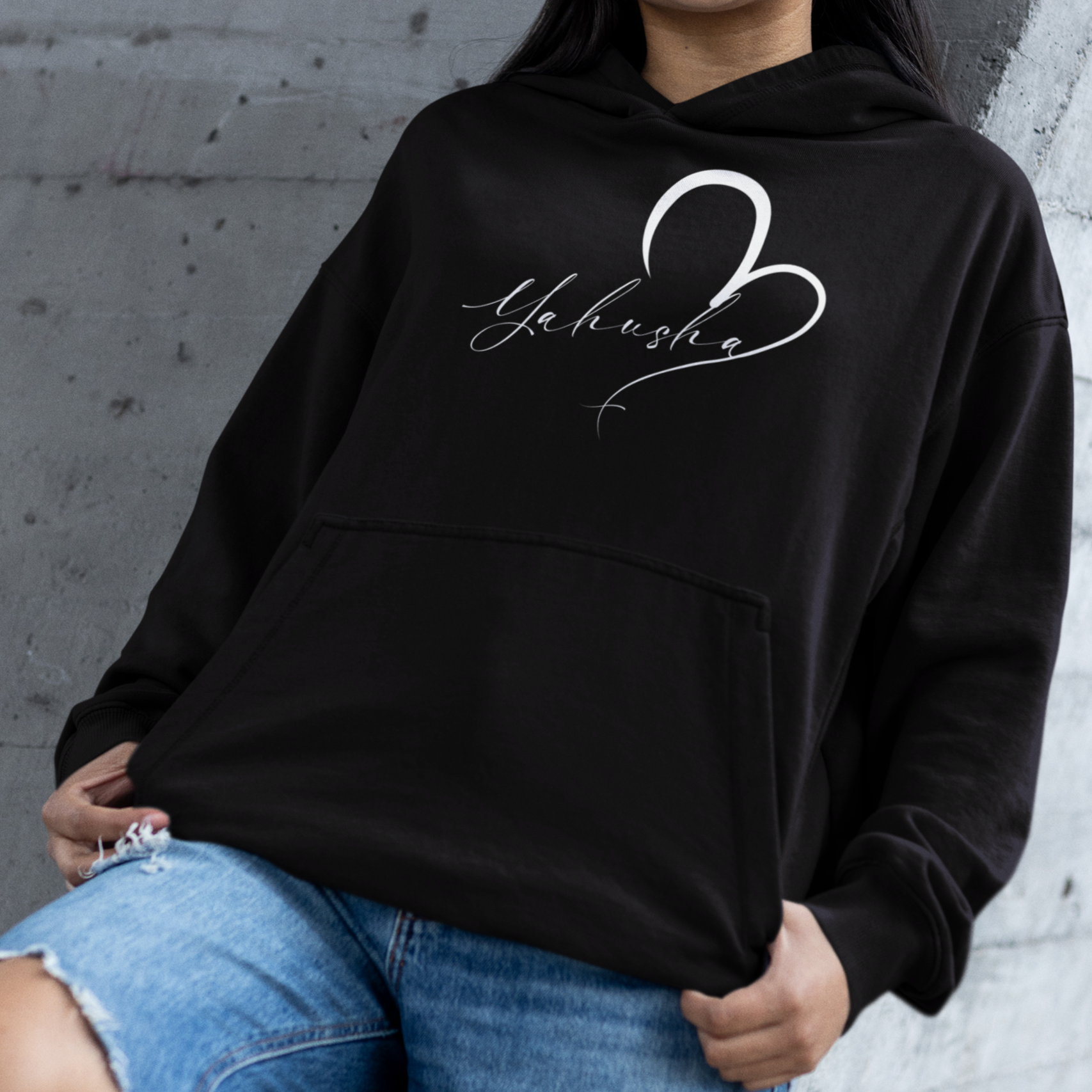 Person wearing a black hoodie with a heart design and text, standing against a concrete wall.