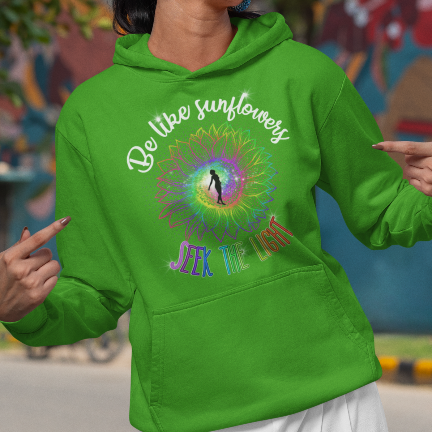 Woman wearing a green hoodie with a colorful design and text, standing in front of a mural.