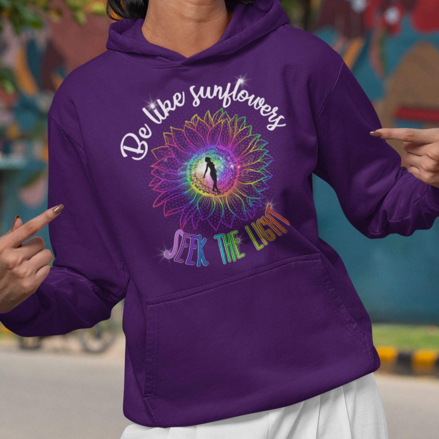Woman wearing a purple hoodie with a colorful design and text, standing in front of a mural.